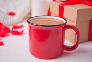 Kırmızı kahve kupası, hediye kutusu ve gri masada kırmızı gül yaprakları.