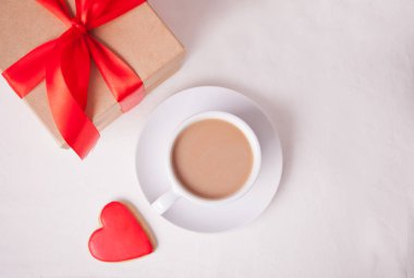 Bir fincan kahve ve beyaz masada hediye kutusu olan kalp şeklinde kırmızı kurabiye.