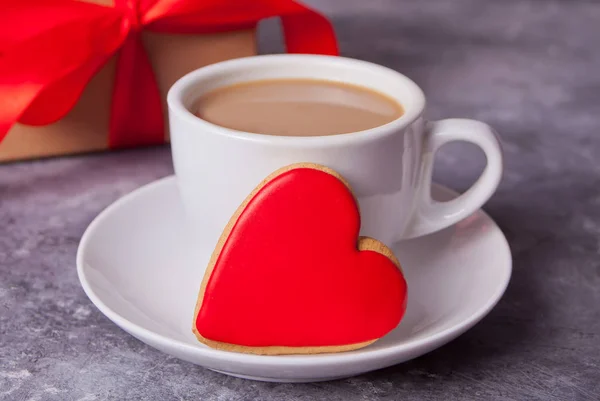 Cup of coffee and a heart shaped red cookie with gift box on the background. — Stock Photo, Image