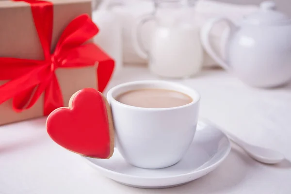 Cup of coffee and a heart shaped red cookie with gift box and teapot on the white table — Stock Photo, Image