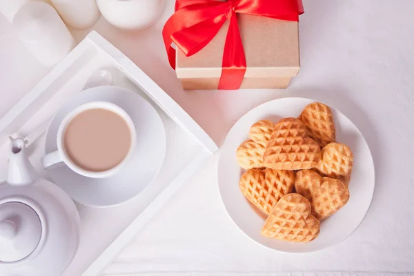 Taza de té con leche, galletas en forma de corazón y caja de regalo en la mesa blanca . — Foto de Stock