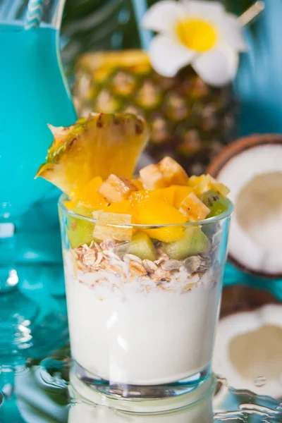 Exotische tropische fruitsalade met muesli en yoghurt in een glas met ananas en kokosnoot op de achtergrond. — Stockfoto