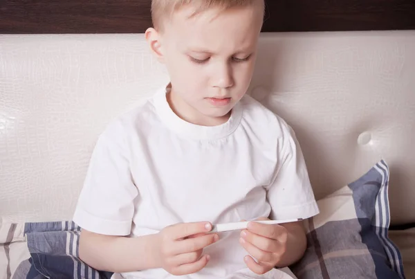 Niño enfermo sentado en la cama y mirando el termómetro digital. El chico malo está midiendo la temperatura corporal y no se siente bien —  Fotos de Stock