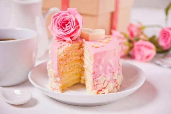 Mini malý dort s růžovou polevou, krásné růže, šálek kávy, dárková krabička na bílém stole. — Stock fotografie