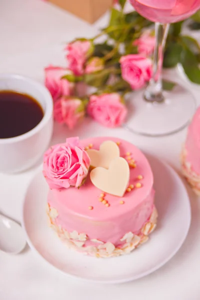 Mini gâteau avec glaçure rose, belles roses, tasse de café, verre de vin rose sur la table blanche . — Photo