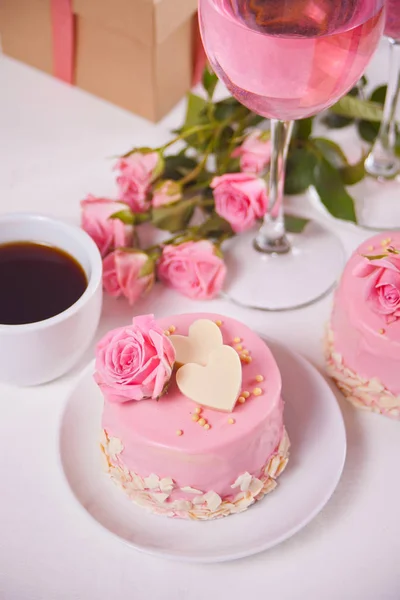 Mini gâteau avec glaçure rose, belles roses, tasse de café, verre de vin rose sur la table blanche . — Photo