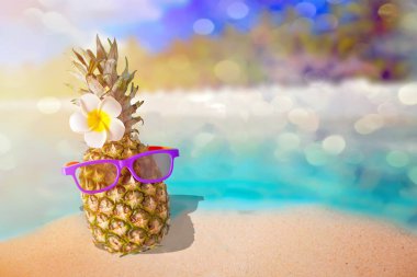 Arkasında deniz ya da okyanus olan beyaz bir kumda güneş gözlüklü, sulu, komik bir ananas. Tatil tropikal kavramı.
