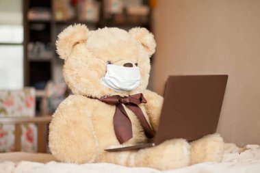 Dijital tablet dizüstü bilgisayarla yatağın üzerinde duran tıbbi maskeli büyük kahverengi ayı oyuncağı. Çevrimiçi arama arkadaşları veya ebeveynler