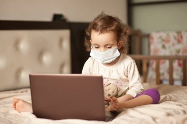 Yatakta oturmuş medikal maskeli şirin bir kız dijital tablet dizüstü bilgisayar kullanıyor. Çevrimiçi arama arkadaşları veya ebeveynler