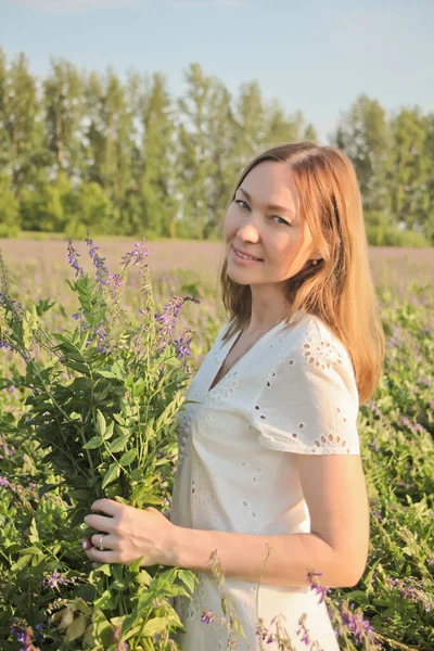 牧草地で白いドレスの若い女性 彼女は手に紫色の花の花束で立っている — ストック写真