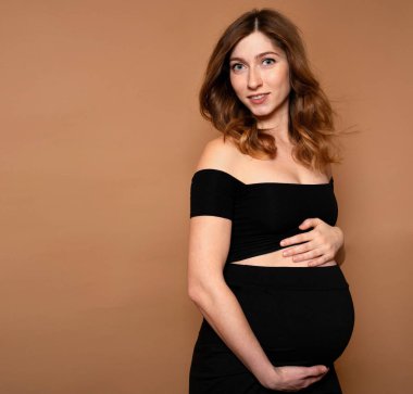 Siyah elbiseli, kahverengi arka planda hamile bir kadının stüdyo çekimi. Annelik kavramı