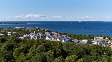 Mavi açık gökyüzü, masmavi denizin üzerinde küçük bir bulut şeridi. Sahilde bir tatil köyü ve bir sürü yeşil ağaç var.