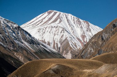 İki kahverengi dağ arasında beyaz, karla kaplı bir dağ zirvesi. Açık mavi gökyüzü ile güneşli bir gün. Merkezde bir dağ yaylası var..