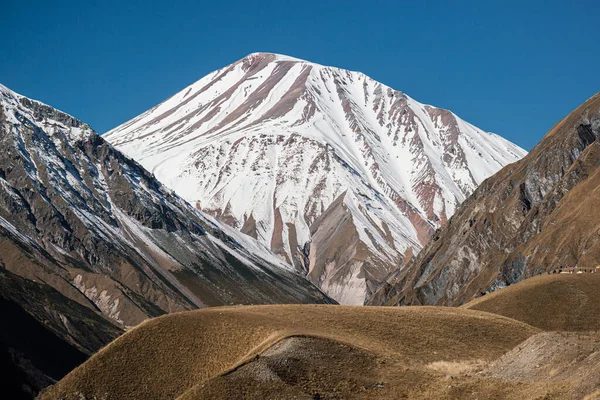 İki kahverengi dağ arasında beyaz, karla kaplı bir dağ zirvesi. Açık mavi gökyüzü ile güneşli bir gün. Merkezde bir dağ yaylası var..