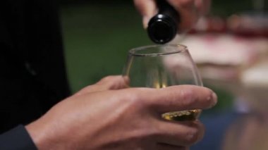 Erkek elleri yeşil bir cam şişeden temiz bir bardağa beyaz şarap döker, sonra da şişeyi masanın üzerine koyar.