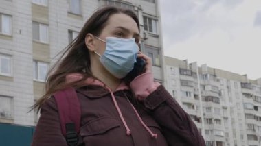 Uzun saçlı, yüzünde tıbbi maske olan beyaz bir kız Coronavirus salgını sırasında apartmanın arka planında telefonla konuşuyor.