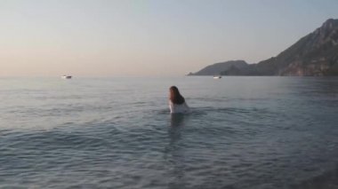 Tatildeki bikinili kadın sahilde suya doğru yürüyor. Deniz suyunda beyaz ıslak bluz giyen güzel genç bir kadın. Mutlu kadın turist gülümsemesi günbatımında sıcak dalgaların tadını çıkarıyor.