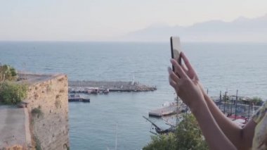 Genç bir kadın akıllı telefondan fotoğraf çekiyor. Eski Antalya, Kaleici limanı, deniz manzarası ve sisin içindeki dağlar.