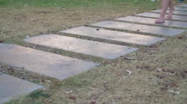Genç bayan bronzlaşmış bacaklar taştan yapılmış yol boyunca gider, yeşil çimenler etraftadır. Çıplak ayakla yan tarafa git, arkanı dön, yerinde dön ve ayağa kalk.