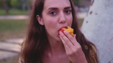 Uzun kahverengi saçlı, sarı elbiseli, bronz tenli bir kadın şeftaliyi ısırıp çiğniyor. Komik kız doğada dinleniyor. Kameraya bakıyor ve yüzler yapıyor, gülümsüyor ve gülüyor. Kapat.