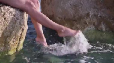 Bronzlaşmış güzel bacaklar deniz suyu sıçratıyor. Genç bir kadın büyük bir taşın üzerinde oturuyor, bacakları şeffaf mavi suda sallanıyor, güneş parlıyor..