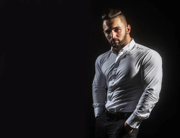 Ciddi adam ellerini onun pantolon ya da pantolon ceplerinde tutar. Tıraşsız genç iş adamı ile spor vücut Beyaz gömlekli. Adam atletik vücut ile kendine güvenen görünümünü. Erkek model siyah izole