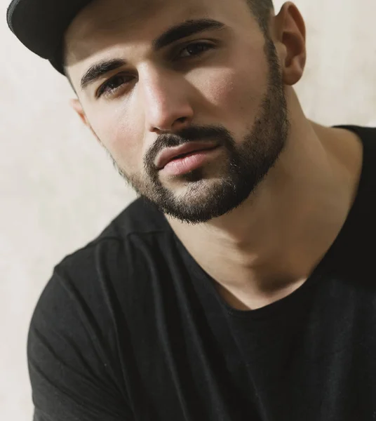 Gorra de hombre en un hombre barbudo blanco. Joven con barba y gorra negra en blanco. Retrato de joven atractivo guapo en una camiseta negra con gorra negra. Hombre mirada seductora sobre un fondo gris — Foto de Stock