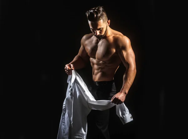 Man undresses, takes off his white shirt. Young guy with beautiful bare naked sports torso. Man wearing shirt. Sexy young  muscular man in shirt on a dark background at morning — Stock Photo, Image