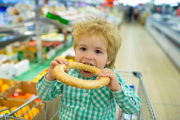 Loja de comida de compras, menino bonito sentado no carrinho de compras no supermercado em compras familiares. Uma mercearia. Criança come pão saboroso na padaria, descontos após o almoço, doces frescos — Fotografia de Stock