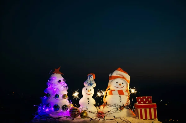 Noche de Navidad, celebración cerca del árbol de nieve de Navidad. Esculturas de nieve por la noche. Dos muñecos de nieve felices en la noche de Año Nuevo. Concepto de celebración festiva. Decoración de invierno. Los muñecos de nieve sostienen luces de Bengala — Foto de Stock