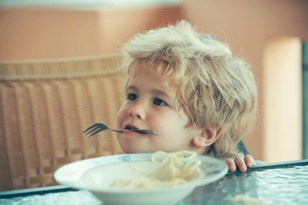 O rapaz engraçado tem garfo nos dentes. Miúdo alegre come esparguete na pizzaria. A criança estraga com comida, comida de bebê como entretenimento ou brincadeira. Prato com comida para bebê na mesa — Fotografia de Stock