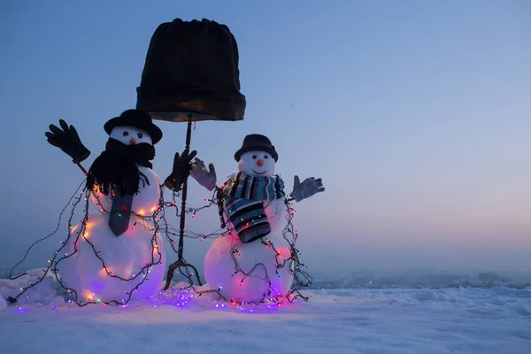 Lindos muñecos de nieve vestidos con ropa oscura de pie en la nieve. Gran lámpara de pie negro detrás de muñecos de nieve. Foto nocturna de instalación de invierno — Foto de Stock