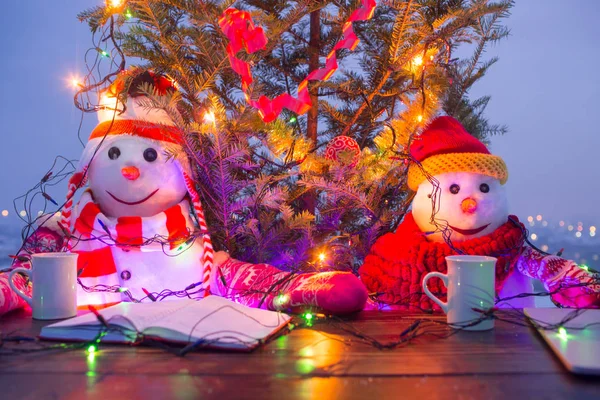 Muñecos de nieve tomando té en la mesa de terraza. El ambiente navideño afuera. Luces brillantes y ambiente acogedor. Bebida caliente en invierno frío. Muñecos de nieve graciosos, árbol de Navidad detrás. Disfruta del humor navideño — Foto de Stock