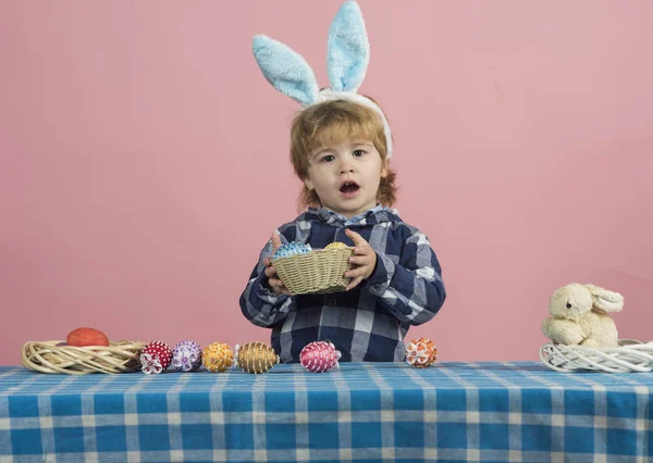 Ostereierschmuck für die Osterfeierlichkeiten. Das süße blonde Kind hält eine Schale mit Ostereiern in der Hand und blickt überraschend in die Kamera. Hasenohren, Osterhasen. gemütliches Zuhause auf rosa Hintergrund, Spielzeug — Stockfoto