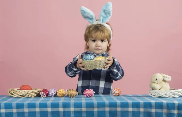 Osterkorb mit Eiern. Junge in blauem Hemd und flauschigen großen Ohren zeigt große Schale voller Ostereier auf rosa Hintergrund. Ostereier mit kleinem Sohn auf Meisterkurs verziert liegen als Dekoration auf dem Tisch — Stockfoto