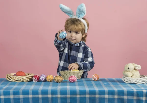 Osterei, niedlicher blonder Junge mit braunen Augen zeigt Spielzeugei aus Schale. Tisch mit blau-weißer Tischdecke, Ostereiern, Osterhasen und Schalen aus Stroh. Urlaubstradtionskonzept — Stockfoto