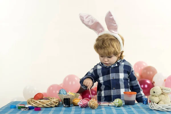 Junge malt Ostereier zu Hause am Tisch im gemütlichen Wohnzimmer. Familientradition und Kreativität. rosa Hasenohren auf dem Kopf eines hübschen Jungen. Vorbereitung auf Ostern — Stockfoto