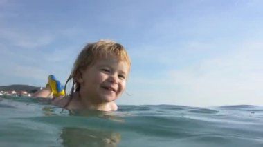 Mutlu çocuk gülüyor ve denizde yüzüyor. Alt yüzündeki gülümseme. Bebek anne ile plajda okyanusta yüzmek öğrenmektir. Çocuklar, turizm ve mutlu sağlıklı çocukluk ile yaz tatili. Koca çocuk gülümseme