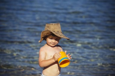 Küçük şirin çocuk sahil üzerinde duruyor ve sarı plastik kova tutar. Arka plan ve büyük kahverengi şapkalı çocuk açık deniz fotoğraf makinesi vasıl görünüyor. Aile tatilleri ve sahil üzerinde tüyler ürpertici. Kovboy sahilde