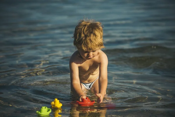 金髪の少年は、水の 3 つのボートを果たしています。子供が水で遊ぶの穏やかな、暖かい一日。海岸で子供のためのゲーム。子供のためのレクリエーションの休日。ボートで遊ぶに焦点を当てた子 — ストック写真