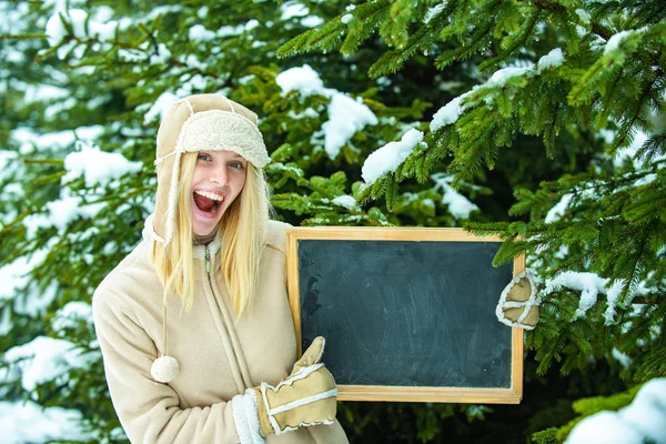 Cool advertisement for the winter. A girl in a warm hat and gloves holds an announcement board. Thumb up from a young woman on the background of a winter forest. Christmas tree.