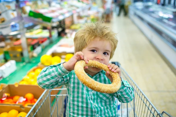 Criança esfomeada. Um rapaz giro come um pão no supermercado. Lanche para uma criança enquanto faz compras . — Fotografia de Stock