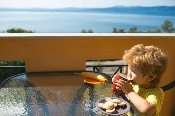隔離中に自宅で朝食。自己分離。コロナウイルスと子供の健康。健康的な朝食。海のそばのテラスで牛乳を飲む子供. — ストック写真