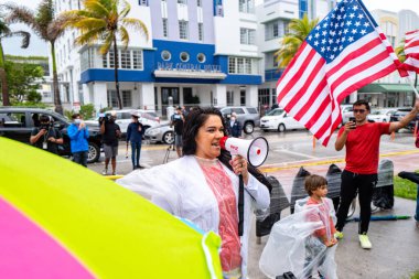 Miami Beach, Florida, ABD - 10 Mayıs 2020: Coronavirus gösterisi. Amerikan bayrağının arka planına karşı hoparlörlü cesur Miami kadını. Güney Florida sakinleri Covid 19 yasağına karşı.