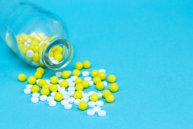 Cam kavanozdan beyaz ve sarı haplar döküldü. Kopyalama alanı olan mavi arkaplan üzerindeki ilaç kapsülleri.