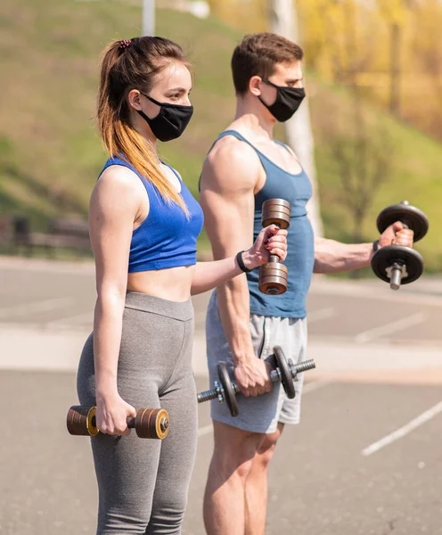Athletic guy and girl doing exercises with dumbbells on the sports field in medical masks. COVID-19. Health care.