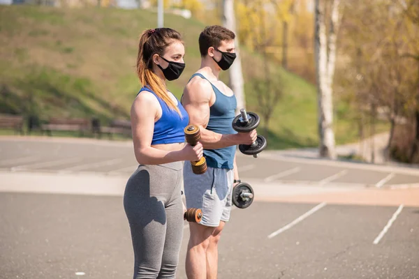 Athletic guy and girl doing exercises with dumbbells on the sports field in medical masks. COVID-19. Health care.