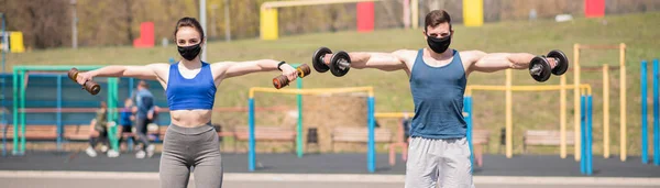 Athletic guy and girl doing exercises with dumbbells on the sports field in medical masks. COVID-19. Health care.