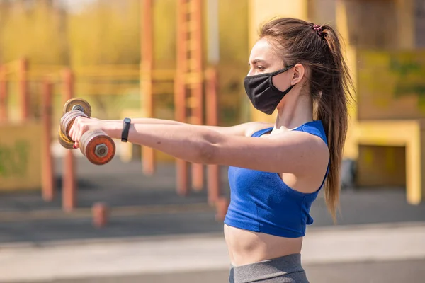 Young athletic girl doing exercises with dumbbells in a medical mask on the playground during a pandemic. COVID-19. Health care.