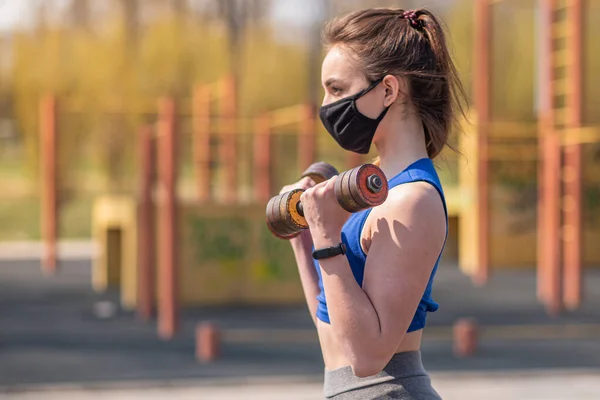 Young athletic girl doing exercises with dumbbells in a medical mask on the playground during a pandemic. COVID-19. Health care.